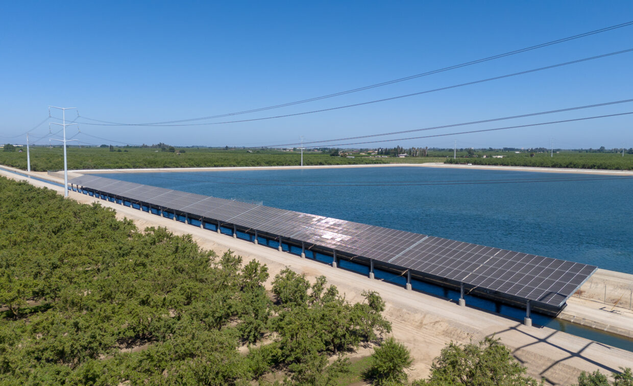 Aerial photo of solar panels over irrigation canal with reservoir in background.