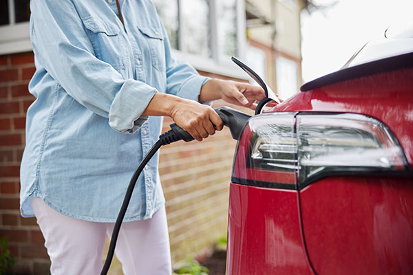 Woman attaches charging cable to red electric vehicle outside her home.