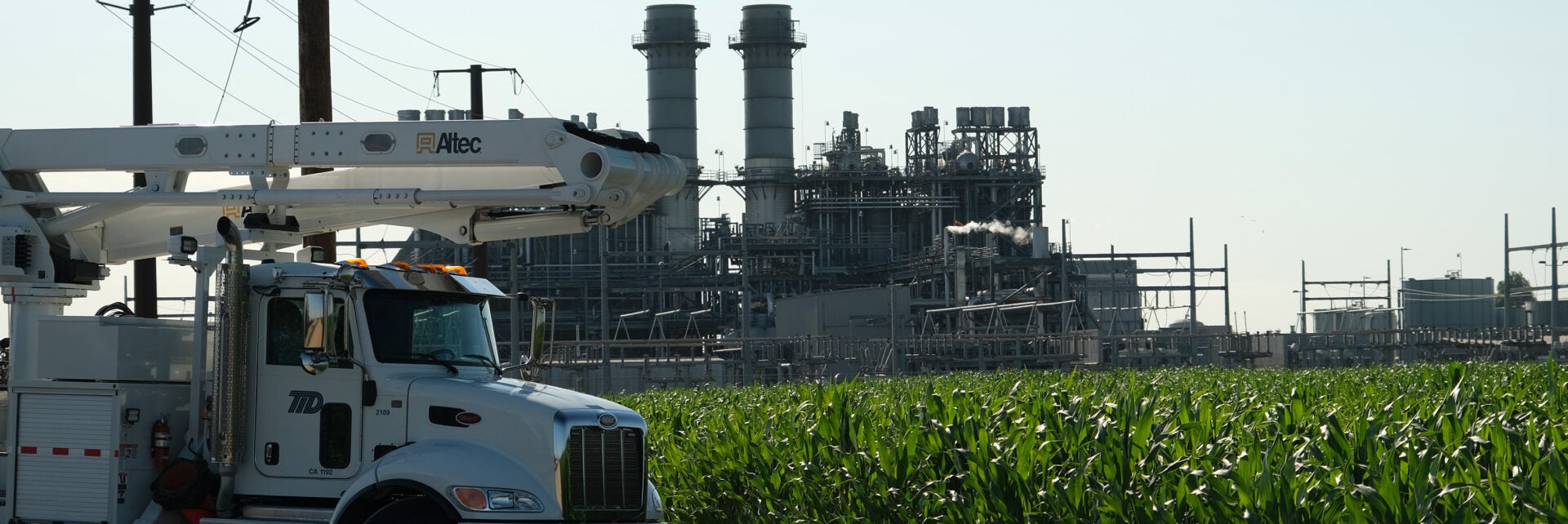 Bucket truck with power plant and corn field in the background.